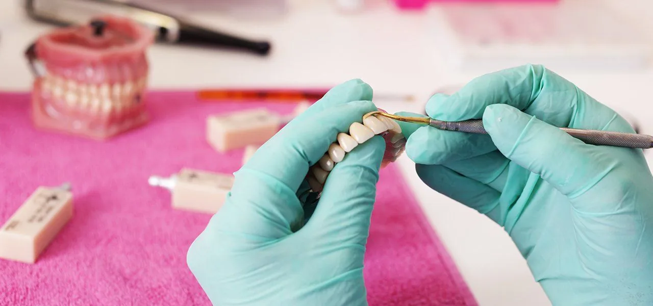 Gloved hands working on a row of denture teeth with a fine metal instrument